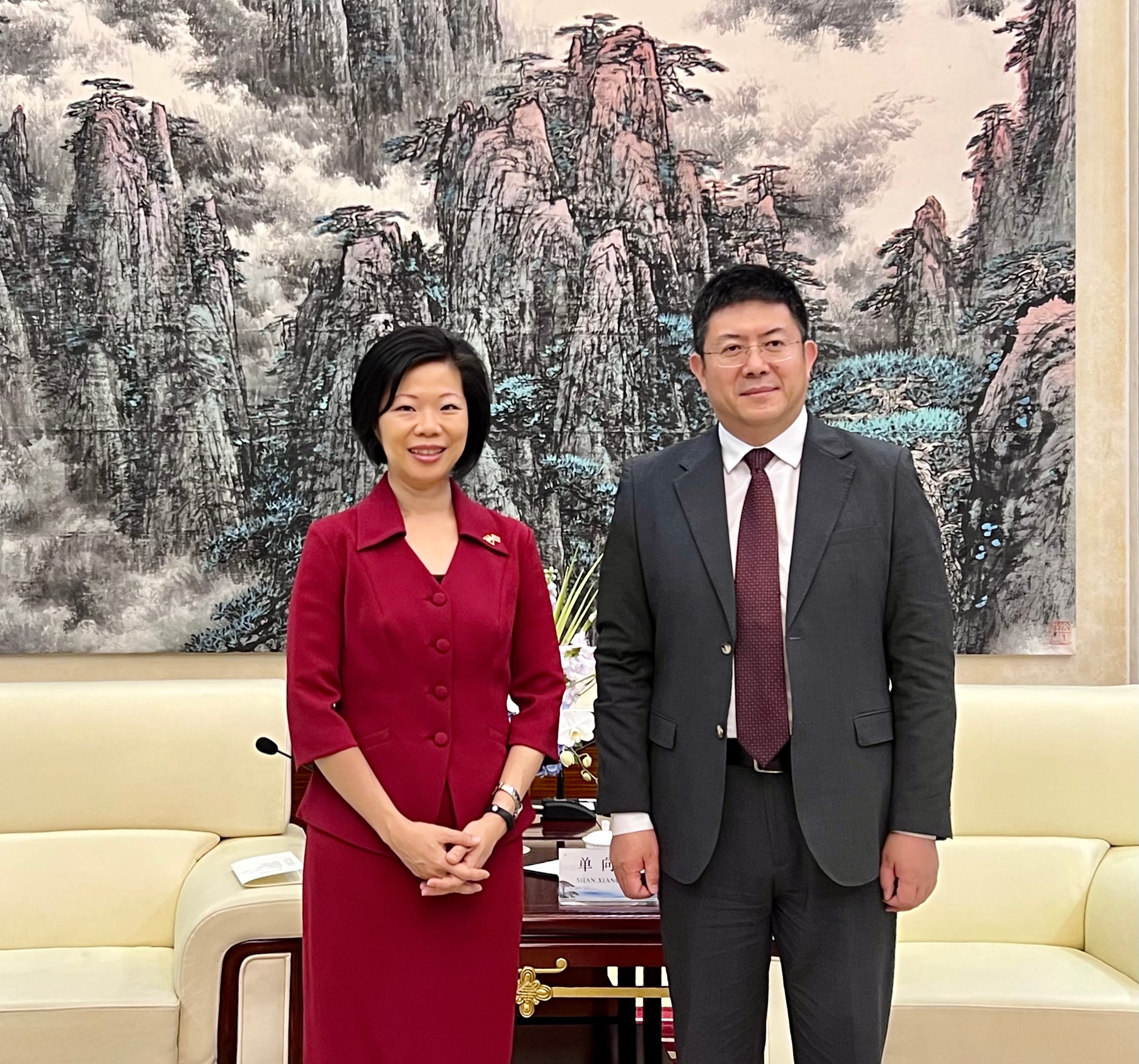 Two people pose in front of a mountainscape painting. The woman wears a red suit; the man wears a dark gray suit.